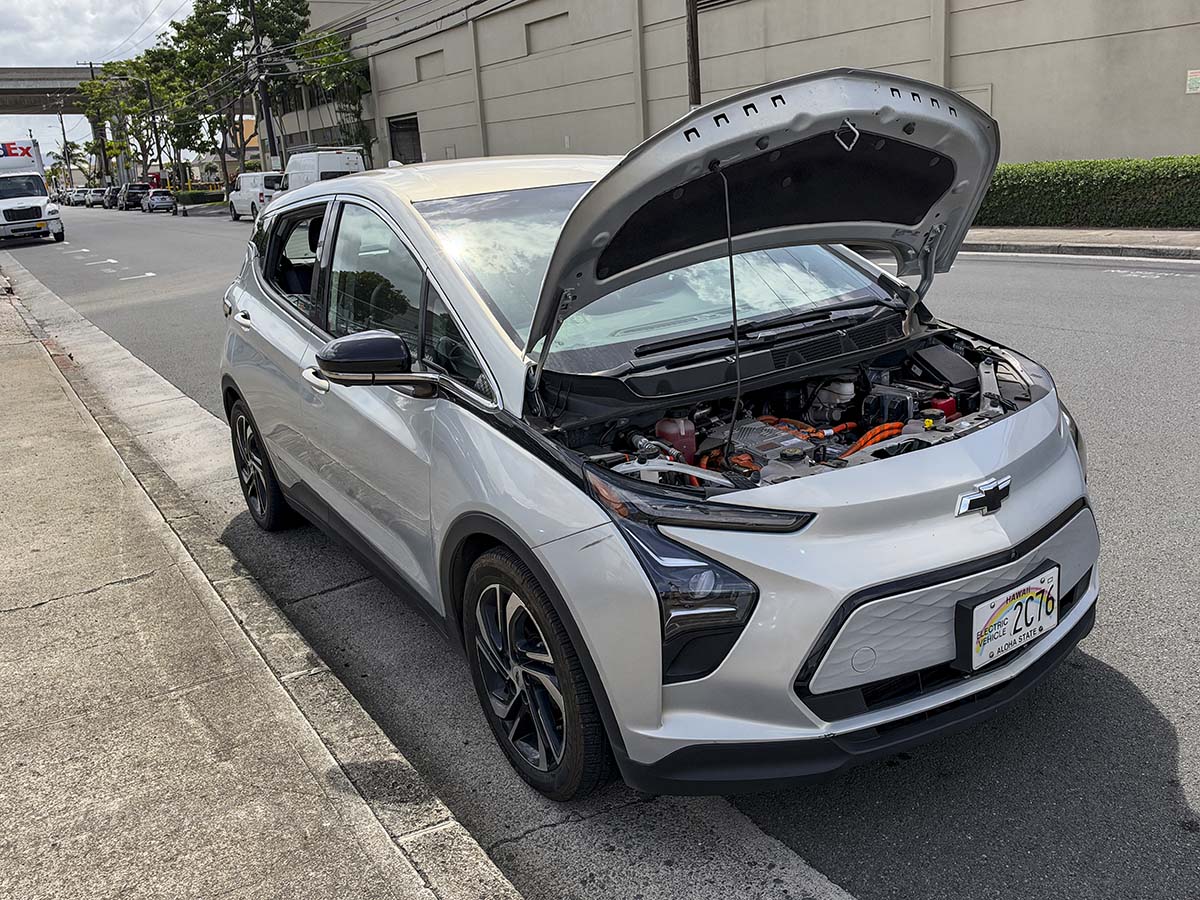 Silver 2024 Chevy Bolt parked along the side of a busy street with the hood up showing the electric motor